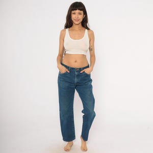May include: A woman wearing a cream-colored tank top and blue denim jeans. The jeans have a straight leg fit. The woman is standing in front of a white background.