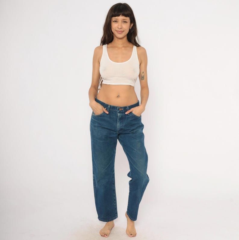 May include: A woman wearing a cream-colored tank top and blue denim jeans. The jeans have a straight leg fit. The woman is standing in front of a white background.