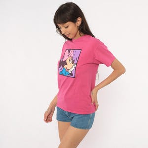 May include: A bright pink short-sleeved t-shirt featuring a square graphic print on the chest. The graphic depicts a cartoon mouse character with a pink bow, wearing a blue top, set against a light purple background with a dark border.