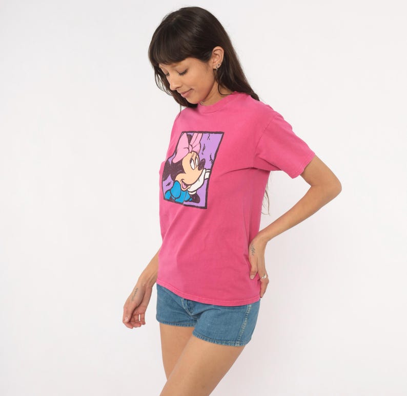 May include: A bright pink short-sleeved t-shirt featuring a square graphic print on the chest. The graphic depicts a cartoon mouse character with a pink bow, wearing a blue top, set against a light purple background with a dark border.