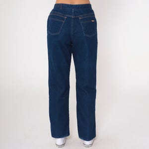 K&ouml;nnte beinhalten: Dunkelblaue Jeans mit geradem Bein. Die Jeans haben ein klassisches F&uuml;nf-Taschen-Design, kupferfarbene N&auml;hte und einen kleinen Logoaufn&auml;her auf der Ges&auml;&szlig;tasche. Getragen mit wei&szlig;en Turnschuhen.