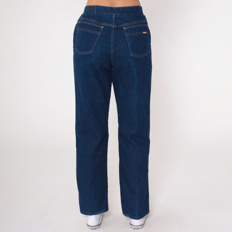 K&ouml;nnte beinhalten: Dunkelblaue Jeans mit geradem Bein. Die Jeans haben ein klassisches F&uuml;nf-Taschen-Design, kupferfarbene N&auml;hte und einen kleinen Logoaufn&auml;her auf der Ges&auml;&szlig;tasche. Getragen mit wei&szlig;en Turnschuhen.