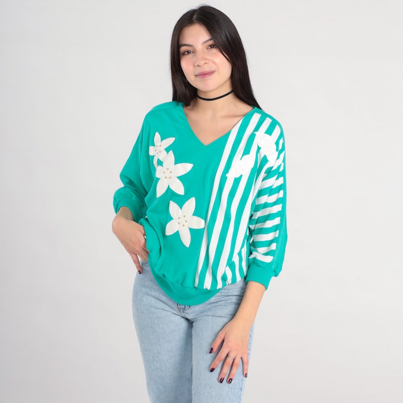 May include: A turquoise and white striped long-sleeved shirt with white flower appliques. The shirt has a v-neck and a relaxed fit.
