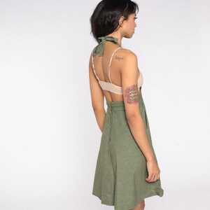 May include: Olive green halter dress with a matching neck tie. The dress features a fitted bodice and a flared skirt. The back of the dress has a zipper closure. The model is wearing a nude-colored bra.