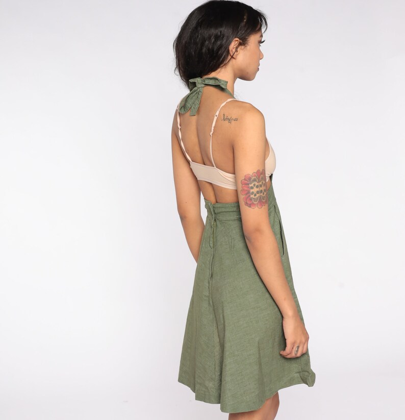 May include: Olive green halter dress with a matching neck tie. The dress features a fitted bodice and a flared skirt. The back of the dress has a zipper closure. The model is wearing a nude-colored bra.