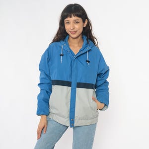 May include: A blue, gray, and navy color-blocked jacket with a hood. The jacket has a zipper closure and two front pockets. The model is wearing the jacket with blue jeans.