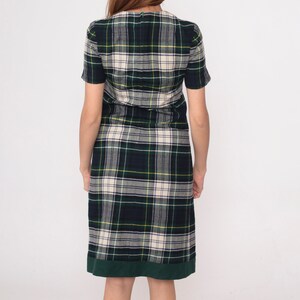 May include: A short-sleeved plaid dress in green, navy, cream, and yellow. The dress has a green hem and a V-neck. The fabric appears to be a woven material. The dress is knee-length and has a classic, timeless style.