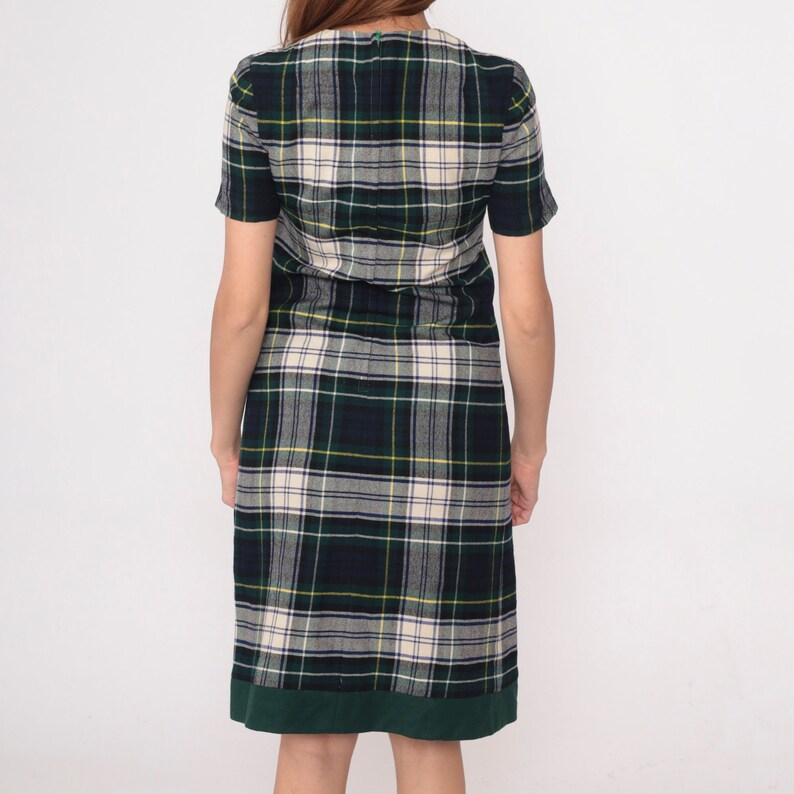 May include: A short-sleeved plaid dress in green, navy, cream, and yellow. The dress has a green hem and a V-neck. The fabric appears to be a woven material. The dress is knee-length and has a classic, timeless style.