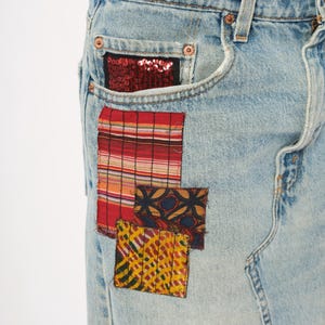 May include: Light blue denim jeans with decorative patches. The patches include a red sequined pocket, a red and white striped patch, and two smaller patches with patterned designs in yellow, red, and blue.