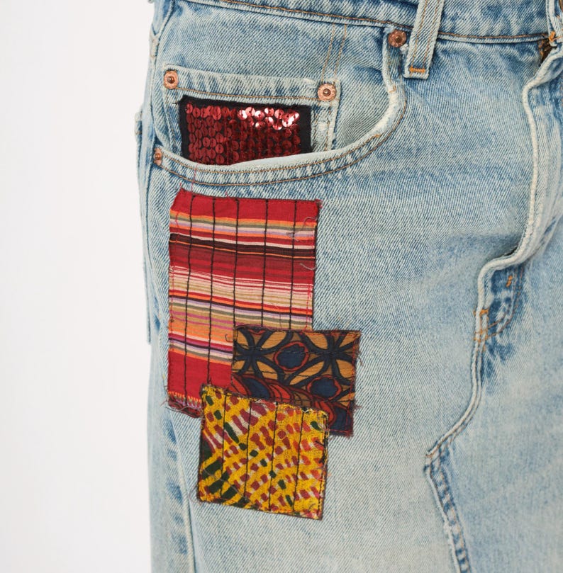 May include: Light blue denim jeans with decorative patches. The patches include a red sequined pocket, a red and white striped patch, and two smaller patches with patterned designs in yellow, red, and blue.