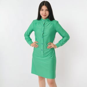 May include: A bright green dress with long sleeves and a button-down front. The dress has a mandarin collar and a straight, knee-length skirt. The model is wearing the dress and has her hands on her hips.