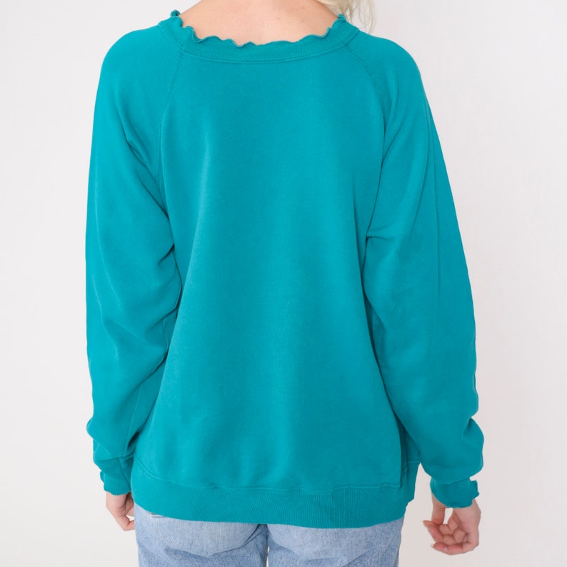 May include: A teal blue sweatshirt with a ruffled neckline. The sweatshirt has long sleeves and a relaxed fit.