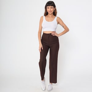 May include: A person wearing a white tank top and brown high-waisted pants. The pants have a straight leg design. The person is wearing white sneakers. The background is plain white.