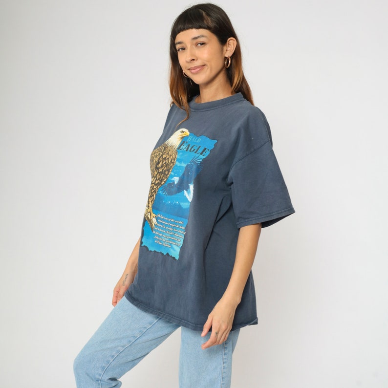 May include: A dark blue t-shirt featuring a graphic of a bald eagle with the words "Bald Eagle" printed on the shirt. The eagle is perched on a branch with a mountain range in the background.