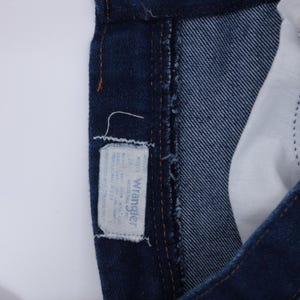 K&ouml;nnte beinhalten: Nahaufnahme einer dunkelblauen Jeans mit einem wei&szlig;en Wrangler-Etikett. Das Etikett zeigt den Markennamen und Gr&ouml;&szlig;eninformationen. Die Jeans hat ein klassisches F&uuml;nf-Taschen-Design und besteht aus strapazierf&auml;higem Denim-Stoff.