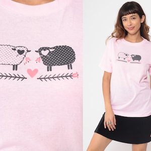 May include: A light pink t-shirt featuring a graphic of two sheep with heart-shaped details, facing each other. One sheep is white with black polka dots, and the other is black with white polka dots. A pink heart is between them.