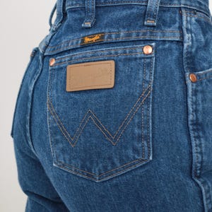 May include: A pair of blue denim jeans with a back pocket featuring a brown leather patch with the Wrangler logo. The jeans have a classic five-pocket style.