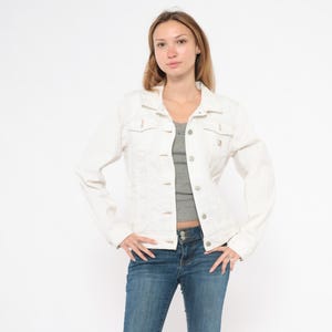 May include: A white denim jacket with button closures, chest pockets, and long sleeves. The jacket is paired with a gray top and blue jeans. The model is standing in front of a white background.