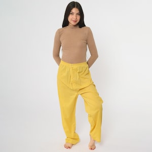 Può includere: Una persona indossa una maglia a collo alto a coste beige e pantaloni gialli. I pantaloni hanno una chiusura con bottoni e una vestibilità ampia. L'outfit è ambientato su uno sfondo bianco.