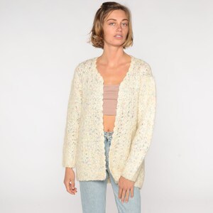 May include: A white cardigan with a scalloped edge and a speckled pattern. The cardigan is open in the front and has long sleeves.