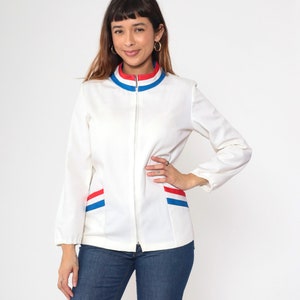 Puede incluir: Una chaqueta blanca con cremallera con ribetes rojos y azules alrededor del cuello y los bolsillos. La chaqueta tiene un cuello alto y mangas largas.