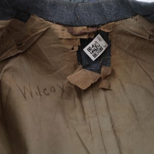 K&ouml;nnte beinhalten: Nahaufnahme des Innenfutters eines grauen Mantels mit zerrissenem Futter. Das Futter ist beige und zeigt das Wort "Wilcox" mit Bleistift geschrieben. Ein Etikett mit einem QR-Code und der Nummer "3831" ist sichtbar.