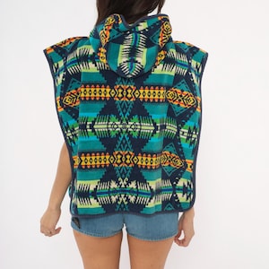 May include: A hooded poncho featuring a dark navy blue base with a vibrant geometric pattern in shades of teal, bright green, yellow, and orange. The sleeveless garment has a hood and a dark navy blue trim along the edges, worn over blue denim shorts.
