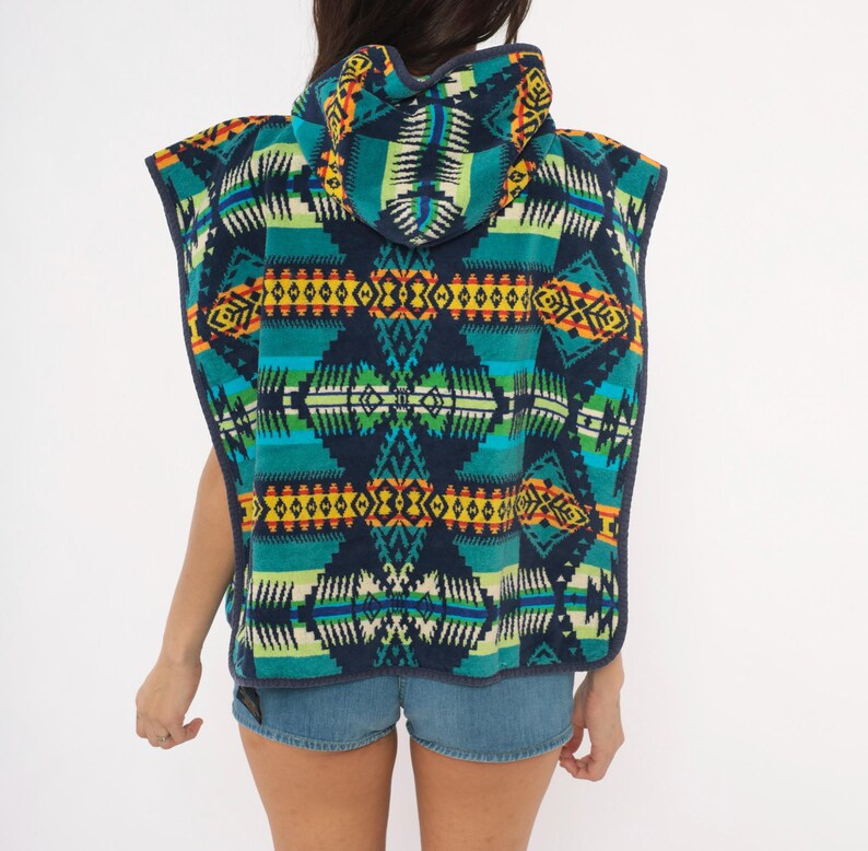 May include: A hooded poncho featuring a dark navy blue base with a vibrant geometric pattern in shades of teal, bright green, yellow, and orange. The sleeveless garment has a hood and a dark navy blue trim along the edges, worn over blue denim shorts.