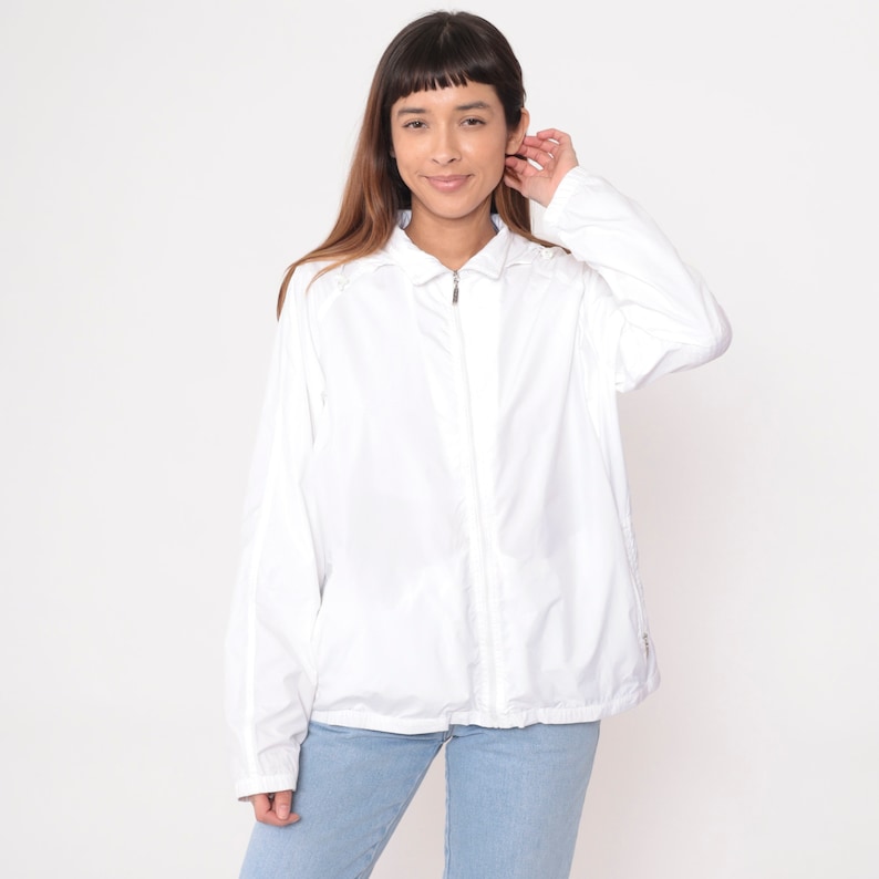 May include: A white, collared jacket with a full-length zipper. The jacket has long sleeves and is worn over light blue jeans. The jacket appears to be made of a lightweight material, suitable for layering. The model is smiling.