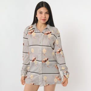 Peut inclure: Une chemise boutonnée gris clair avec un motif de cerf-volant. La chemise à manches longues présente un motif répété de cerfs-volants dans des tons de beige, marron et jaune, avec des rayures horizontales. La chemise a un col et des poignets boutonnés.