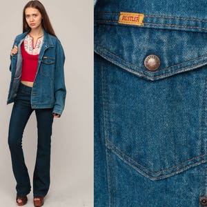 May include: A denim jacket with a buttoned pocket and a "Rustler" label. The jacket is worn over a red top and dark blue jeans. The jeans have a flared leg. The model is wearing brown platform sandals.