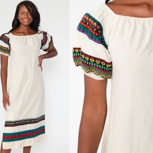 May include: A white off-the-shoulder dress with colorful embroidered trim around the neckline and sleeves. The dress has a relaxed fit and a midi length.