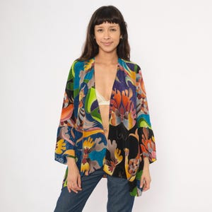 May include: A colorful, open-front kimono-style jacket with a vibrant floral and abstract pattern in shades of blue, orange, purple, and green. The jacket has wide sleeves and is worn over a white bra and blue jeans.