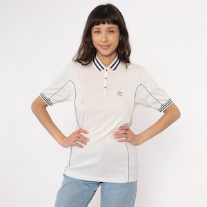 Peut inclure: Polo crème avec garniture bleu marine et petit logo. Le t-shirt a des manches courtes avec des rayures bleu marine et un col. Le t-shirt est boutonné et présente un design classique et sportif.