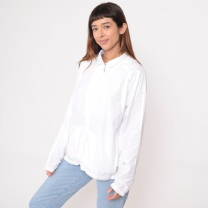 May include: A white, zip-up jacket with a collared neckline. The jacket has long sleeves and a relaxed fit. The model is wearing light blue jeans. The background is plain white.