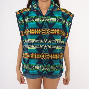 May include: A sleeveless hooded poncho featuring a vibrant geometric pattern in shades of navy blue, teal, bright yellow, and orange. The garment has a V-neckline, wide arm openings, and a repeating symmetrical design across the entire surface.