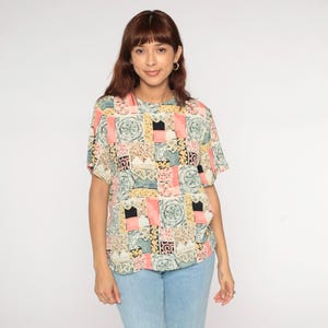 May include: A short-sleeved top with a colorful patchwork design. The pattern includes squares of pink, yellow, black, and teal, with circular and geometric motifs. The top is worn with light blue jeans.