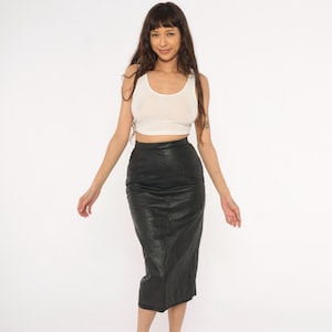 May include: A woman wearing a white tank top and a black leather pencil skirt. The skirt is knee-length and has a textured appearance. The top is a scoop neck style.