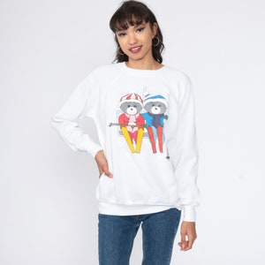 Peut inclure: Sweat-shirt blanc avec un motif de deux ours en dessin animé portant des bonnets d'hiver et skiant en descendant une montagne. Les ours portent des vêtements rouges et bleus.
