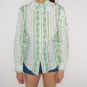 Puede incluir: Una camisa blanca de manga larga con botones y un estampado floral verde. La camisa tiene un cuello puntiagudo y un cierre de botones en la parte delantera.