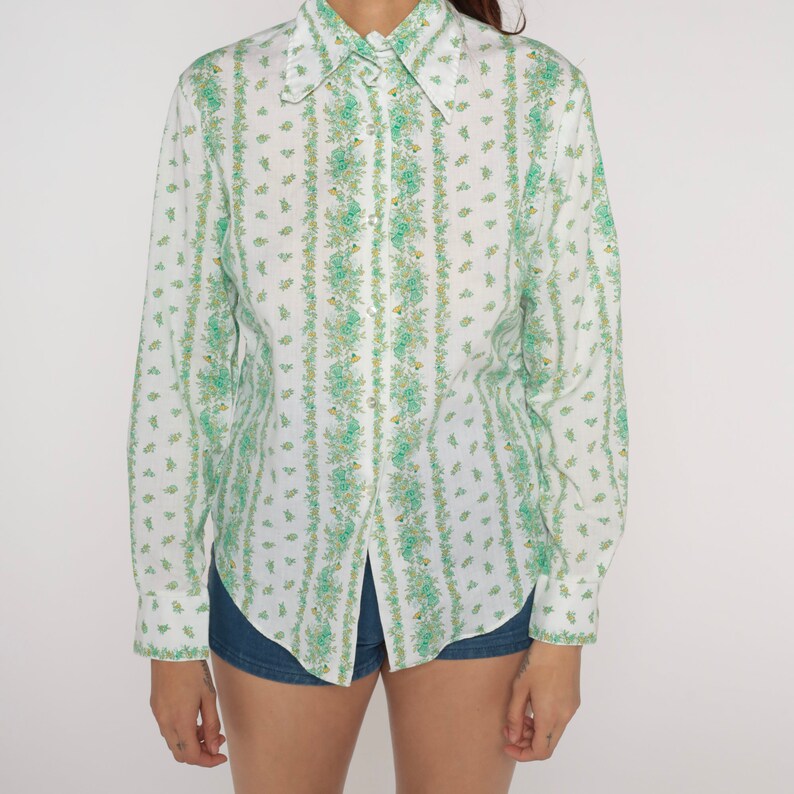 Puede incluir: Una camisa blanca de manga larga con botones y un estampado floral verde. La camisa tiene un cuello puntiagudo y un cierre de botones en la parte delantera.