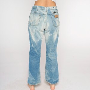 May include: Light blue denim jeans with a faded wash effect. The jeans have a high waist, a Wrangler label on the back pocket, and a slight flare at the hem. The jeans are worn by a person.