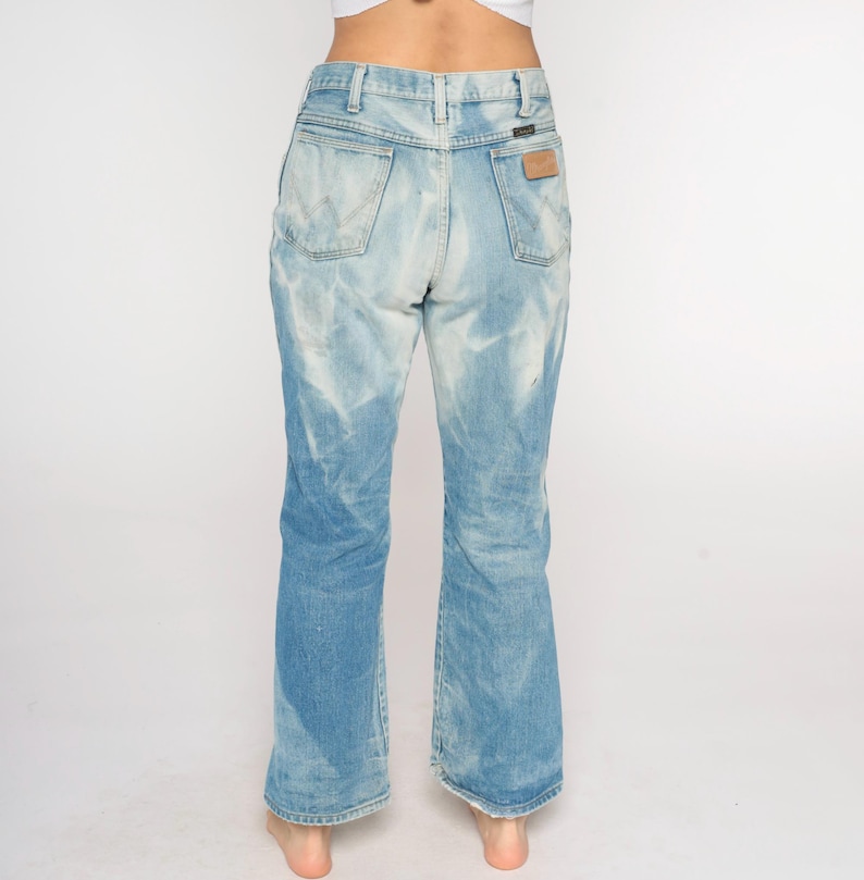 May include: Light blue denim jeans with a faded wash effect. The jeans have a high waist, a Wrangler label on the back pocket, and a slight flare at the hem. The jeans are worn by a person.
