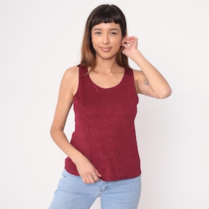 Puede incluir: Un top sin mangas burdeos con cuello redondo. La parte superior tiene una apariencia texturizada y se usa con jeans azul claro. La modelo sonríe y tiene el pelo castaño.