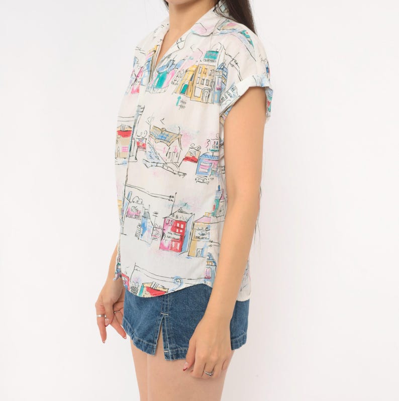 May include: A short-sleeved button-down shirt in white featuring a colorful hand-drawn style print of various buildings and houses in shades of blue, red, yellow, and green, paired with a dark blue denim mini skirt with a front slit detail.
