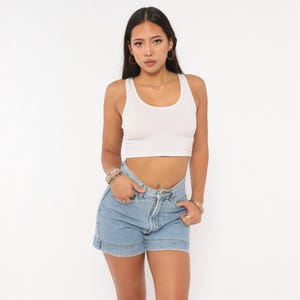 May include: A white sleeveless crop top with a scoop neckline paired with light blue denim shorts. The shorts feature a high-waisted fit, a button fly closure, and a cuffed hemline. The outfit is completed with a stack of beaded bracelets.