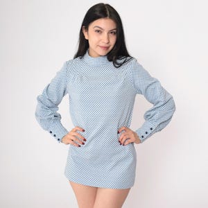 May include: A light blue, long-sleeved top with a high neckline and a pattern of small, dark blue dots. The sleeves have buttoned cuffs. The top is worn by a person.