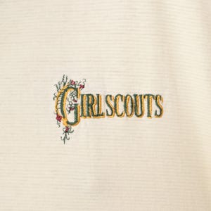 May include: Cream-colored fabric with horizontal stripes, embroidered with the words "GIRL SCOUTS" in a two-tone gold and green font. The text is accompanied by floral embroidery in green, red, and yellow.