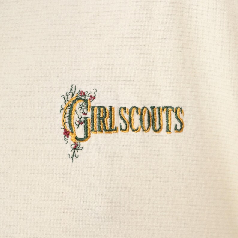 May include: Cream-colored fabric with horizontal stripes, embroidered with the words "GIRL SCOUTS" in a two-tone gold and green font. The text is accompanied by floral embroidery in green, red, and yellow.