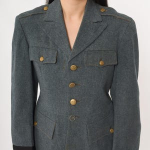 K&ouml;nnte beinhalten: Graue Jacke im Milit&auml;rstil mit goldfarbenen Kn&ouml;pfen und Schulterakzenten. Die Jacke hat zwei Brusttaschen und zwei untere Taschen. Der Kragen ist gekerbt und die &Auml;rmel haben eine dunkle Manschette. Die Jacke ist aus einem strukturierten Stoff.